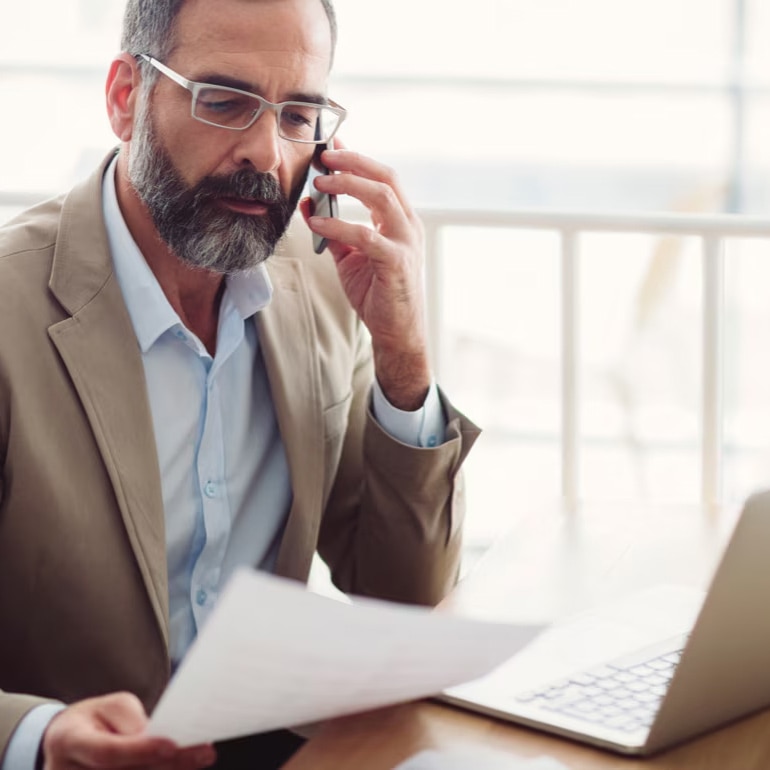 A man with glasses is talking on a smartphone while holding a document and sitting in front of an open laptop.
